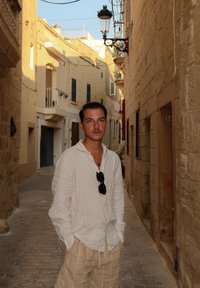 Ozek kameniti ulični prehod s teksturiranimi bež zidovi. Moški v svetli laneni srajci in prugastih hlačah stoji sproščeno, oblečen v sončna očala.