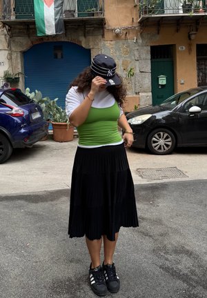 Persona ajustándose una gorra de béisbol negra de pie en una calle urbana, vistiendo una camiseta sin mangas verde sobre una camisa blanca, falda negra plisada y zapatillas negras.