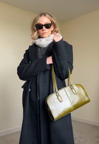 Borsa a mano in pelle verde con due manici superiori, forma rettangolare e texture liscia. Indossata con un cappotto nero e una sciarpa grigia.