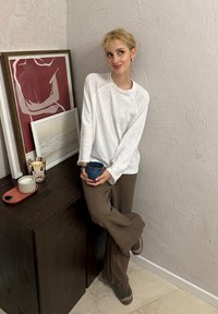 Giovane con capelli biondi corti si appoggia a un armadio di legno scuro, tenendo in mano una tazza blu, accanto a un'opera d'arte incorniciata e candele contro una parete dalla texture.