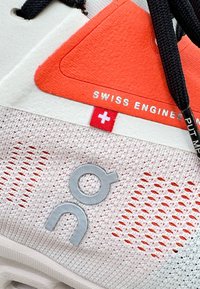 Balti un oranži sporta apavi ar sietveida augšdaļu, gumijas zoli, izteiksmīgu logotipu un teksturētām auduma detaļām. Ietver Šveices karoga akcentu.