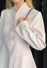 Veste rose clair à manches longues avec une texture lisse, dotée d'un col montant et de poches latérales, tenue d'une main. Design minimaliste.