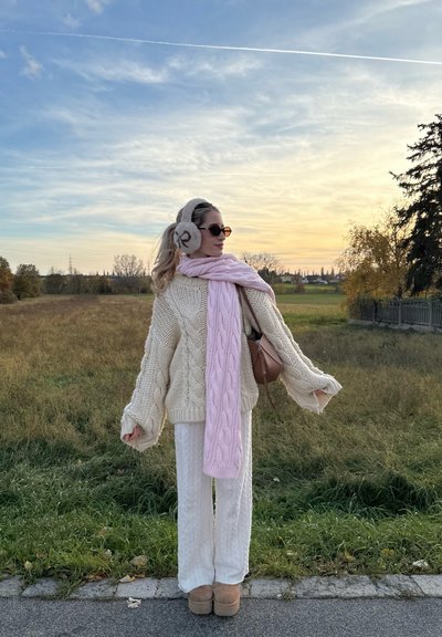Suéter grueso de punto trenzado crema, bufanda de punto trenzado rosa claro, pantalones de pierna ancha blancos, bolso marrón y zapatos de plataforma beige en un campo de hierba.
