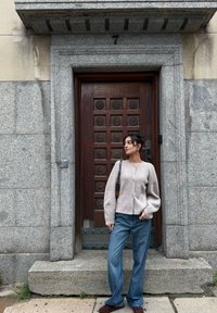 Maglione beige in misto lana con maniche arricciate, bottoni sul davanti e jeans a zampa in denim blu. Scarpe slip-on marrone scuro, in piedi accanto a una porta di legno.