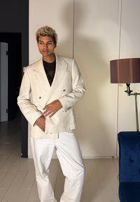 Jeune personne aux cheveux blonds bouclés portant un blazer crème et un pantalon blanc, debout à l'intérieur près d'une lampe sur pied et d'un canapé bleu.