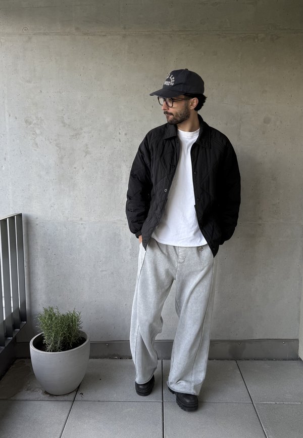 Man wearing black cap, glasses, black jacket, white shirt, and loose gray pants stands on tiled floor near potted plant and concrete wall.