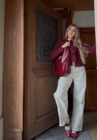 Cardigan croșetat în burgundy intens, combinat cu pantaloni largi albi și pantofi roșii cu toc înalt și baretă unică. Accesorii includ o geantă tote roșie lucioasă.