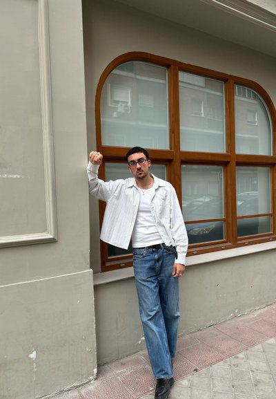 Camisa de botones blanca y gris a rayas sobre una camiseta blanca, jeans azules y zapatos negros. De pie junto a una ventana de madera contra una pared clara.