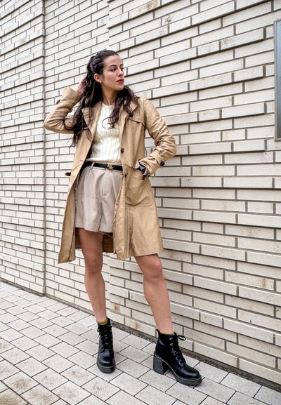 Abrigo trench de cuero marrón claro sobre un suéter de punto blanco, combinado con pantalones cortos beige y botas de tobillo negras con cordones sobre un fondo de azulejos texturizado.