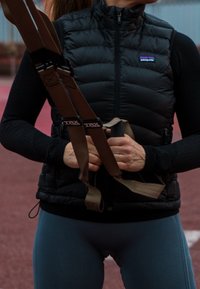 Doudoune sans manches noire avec un col montant, accompagnée d'un haut à manches longues noir. Sangles beiges d'un système d'entraînement en suspension tenues dans les mains.