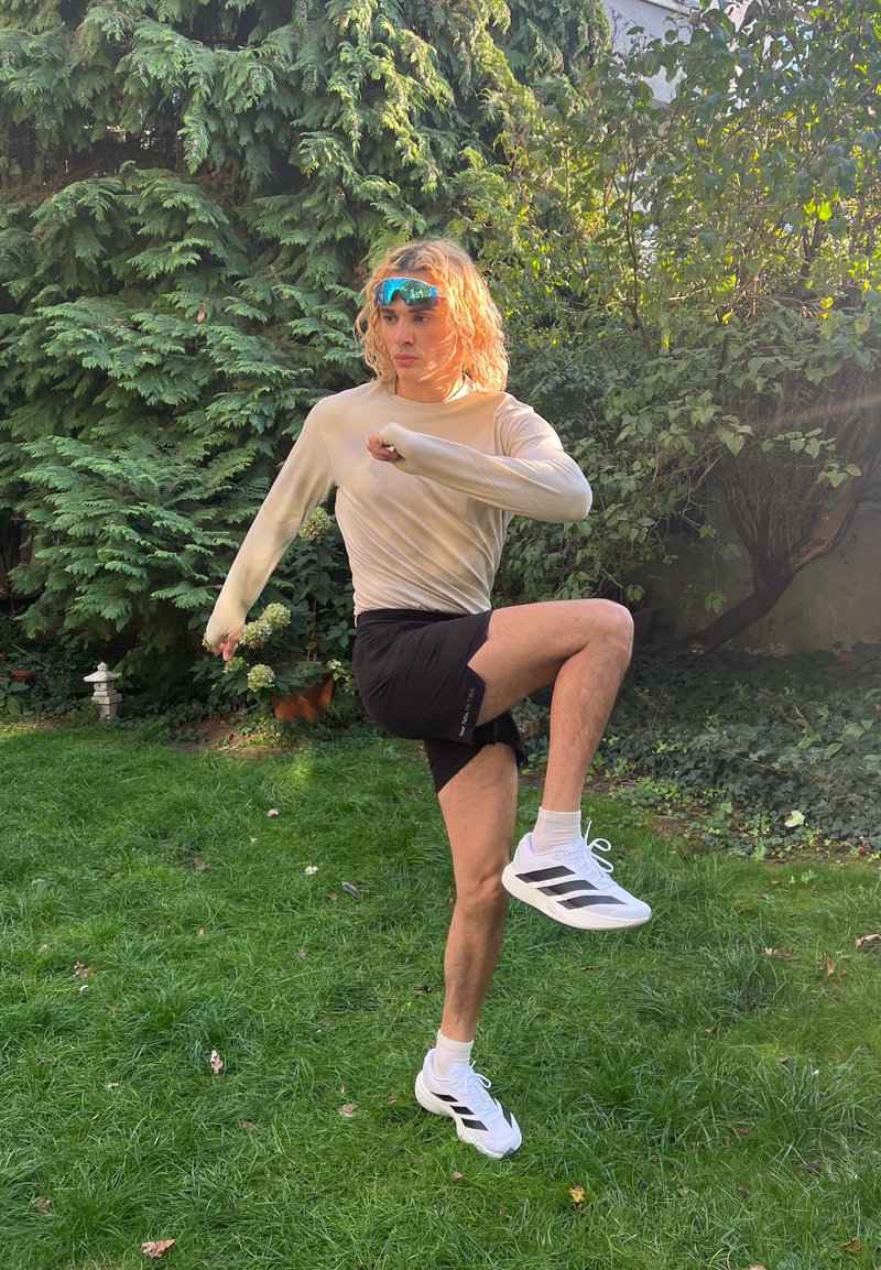 Lange beige shirt, zwarte shorts en witte sportschoenen met zwarte strepen. Staande op gras, voert hij een hoge knie uit.