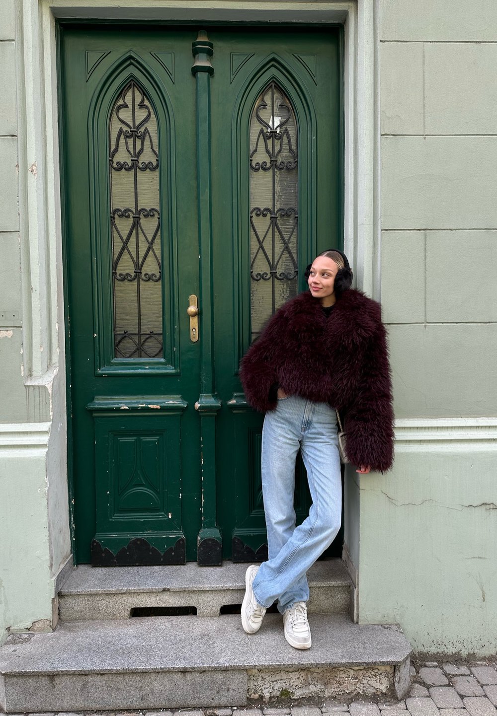 Burgundska umetna krzna jakna, svetlo modri džinsi in bele teniske, stoji poleg zelenih dvojnic z okrasnimi steklenimi in kovinskimi vložki.