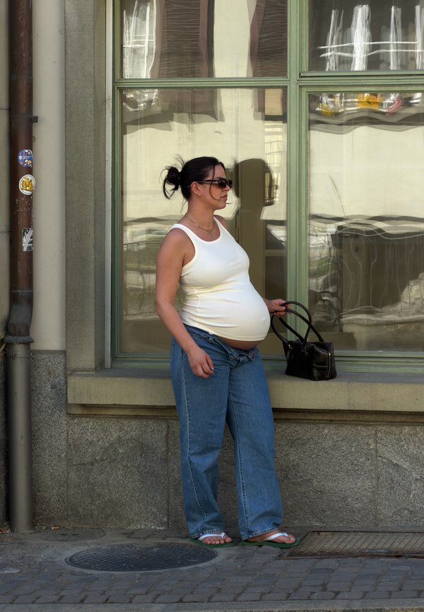Gravid kvinde i hvid tanktop og blå jeans står på fortovet og holder en sort håndtaske med et stort reflekterende vindue bag sig.
