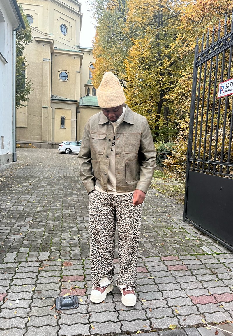 Mann trägt eine beigefarbene Mütze, eine braune Jacke, leopardgemusterte Hosen und weiße Sneaker und steht auf einem gepflasterten Weg mit herbstlichen Bäumen und einem Gebäude im Hintergrund.
