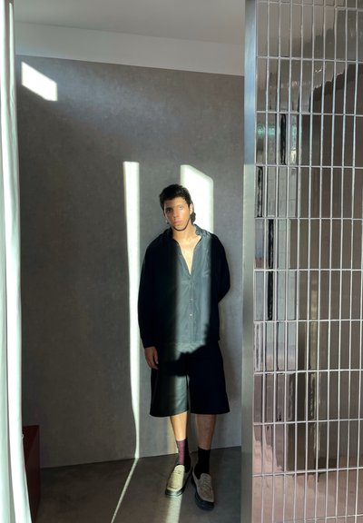 Camisa negra de botones, pantalones cortos negros, zapatos grises; de pie cerca de una pared texturizada y un panel reflectante con patrón de rejilla bajo luz natural.