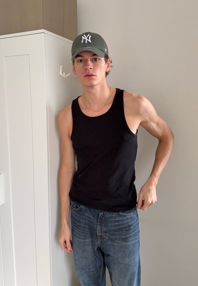 Camiseta negra sin mangas, jeans azules. La camiseta es ajustada, de material acanalado. Se lleva una gorra verde con un logo blanco. El fondo es neutro.