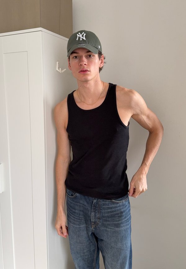 Svart singlet, blå jeans. Singleten er tettsittende i ribbet materiale. En grønn caps med hvit logo bæres. Bakgrunnen er nøytral.