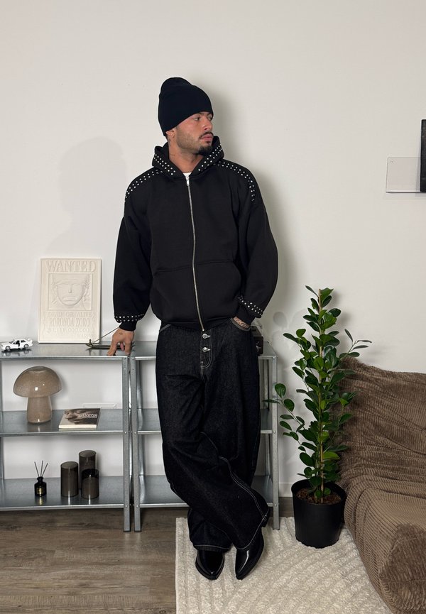 Homme portant un bonnet noir, un sweat à capuche zippé noir clouté et un pantalon large noir, appuyé contre une étagère métallique à côté d'une plante en pot et d'un canapé.