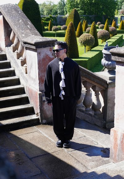 Hombre con atuendo negro con detalles blancos y gafas de sol, de pie cerca de unas escaleras de piedra en un jardín formal con arbustos recortados en forma topiaria.