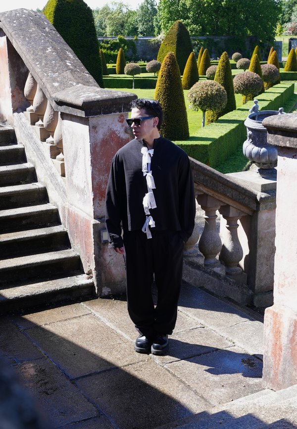 Homme portant une tenue noire avec des accents blancs et des lunettes de soleil, debout près d'escaliers en pierre dans un jardin formel avec des buissons taillés en topiaire.