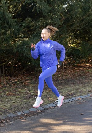Mujer corriendo al aire libre con chaqueta y mallas moradas, calzando zapatillas blancas para correr, con vegetación y un camino de fondo.