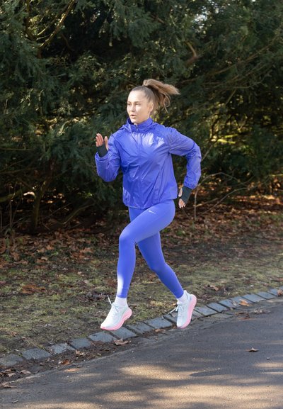 Mujer corriendo al aire libre con chaqueta y mallas moradas, calzando zapatillas blancas para correr, con vegetación y un camino de fondo.