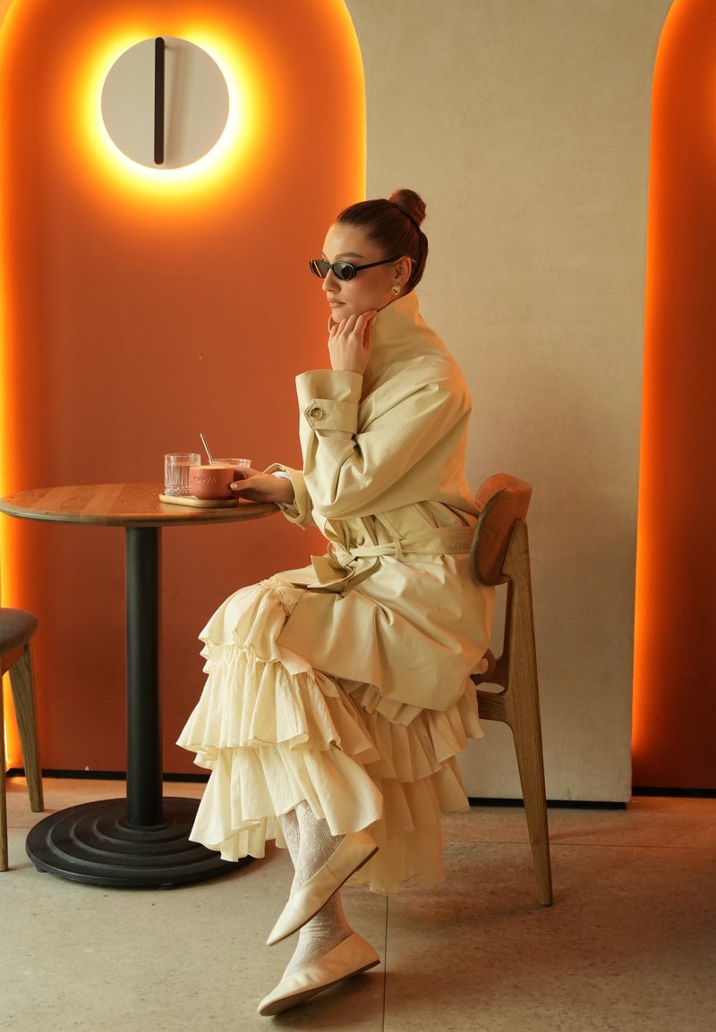 Femme en robe crème superposée et trench-coat assise à une table en bois tenant une tasse, avec une lumière ronde et des panneaux muraux orange derrière elle.