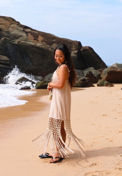 Vestido beige con dobladillo de flecos y detalle de ganchillo. Combinado con chanclas negras en una playa de arena, con olas rompiendo al fondo y acantilados rocosos detrás.
