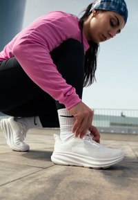 Bílé sportovní boty s výrazným polstrováním, texturovaným povrchem a logem. Osoba má na sobě růžovou dlouhou halenku a černé legíny.
