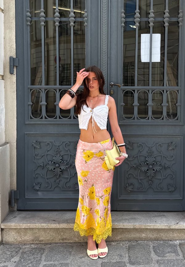 Femme en crop top blanc et jupe rose avec des fleurs jaunes se tient devant un portail en métal gris orné, tenant un sac à main jaune clair.
