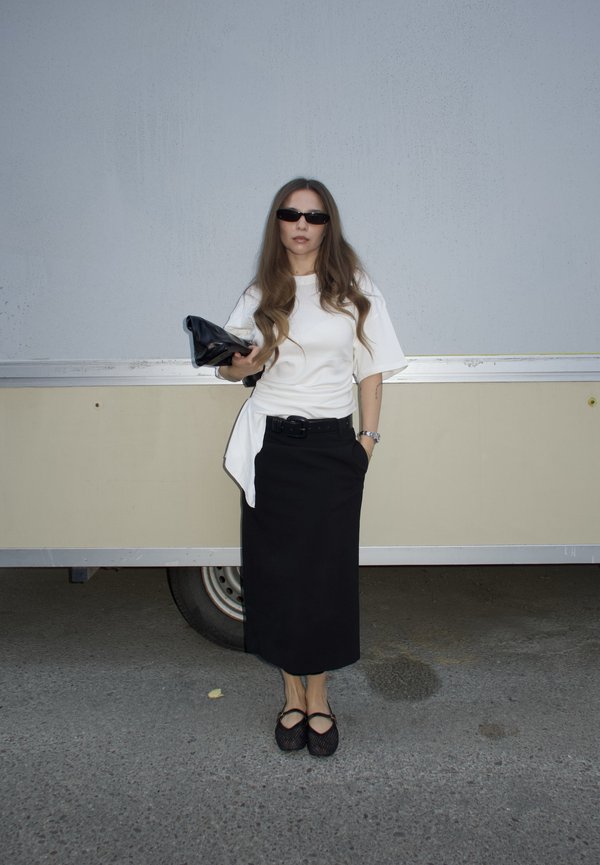Femme portant des lunettes de soleil noires, une chemise blanche oversize, une jupe noire et des chaussures noires, debout tenant une pochette noire devant un véhicule.