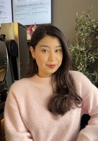 Joven mujer con cabello largo y oscuro que lleva un suéter rosa, está sentada en el interior cerca de una máquina de café y de una planta en maceta verde.