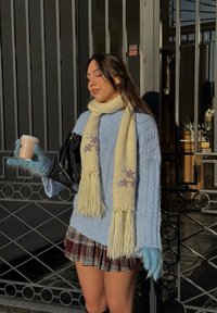 Jeune femme en pull bleu, jupe à carreaux, écharpe et gants, tenant une tasse de café et un sac à main noir, debout près d'un portail métallique sous le soleil.