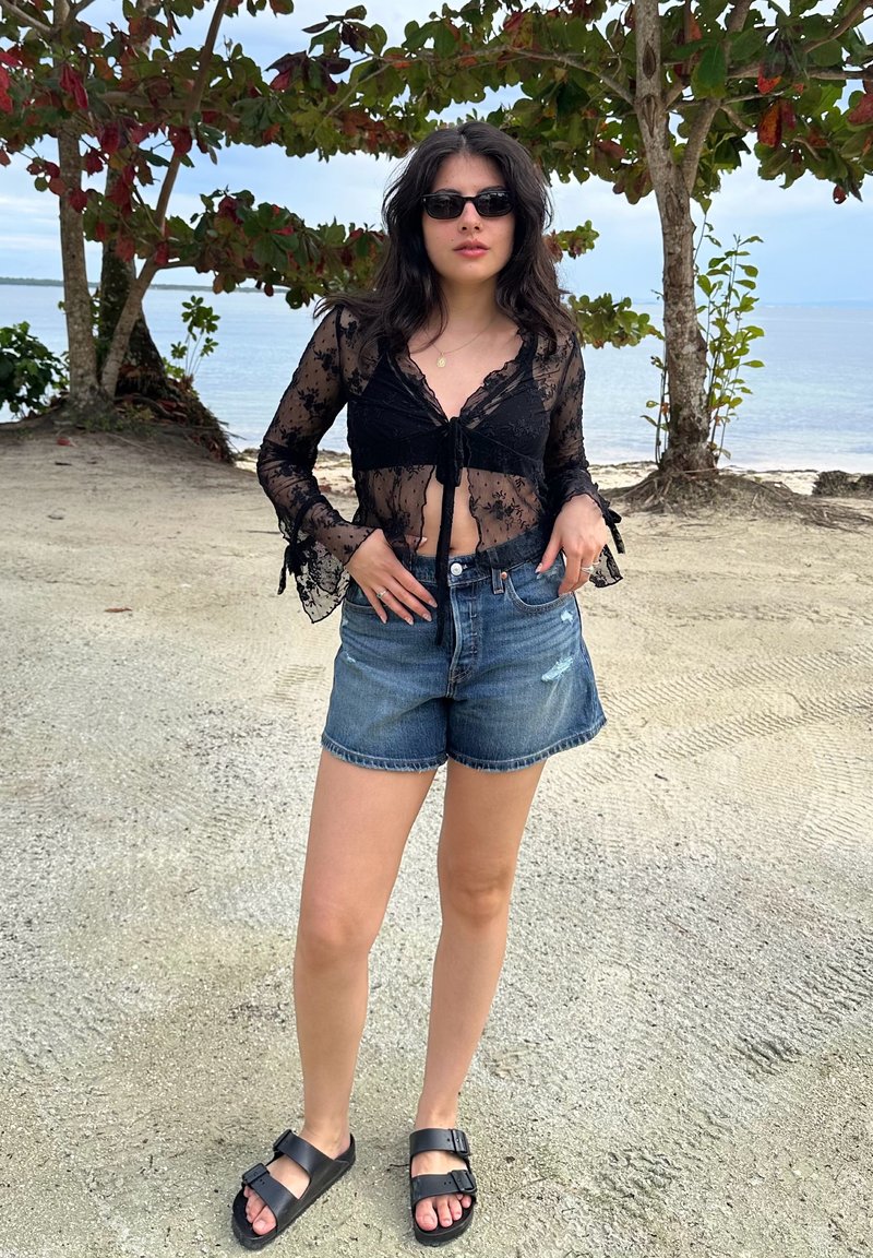 Jeune femme portant des lunettes de soleil noires, un haut en dentelle noire transparente, un short en denim et des sandales noires, debout sur une plage de sable près des arbres et de l'eau.