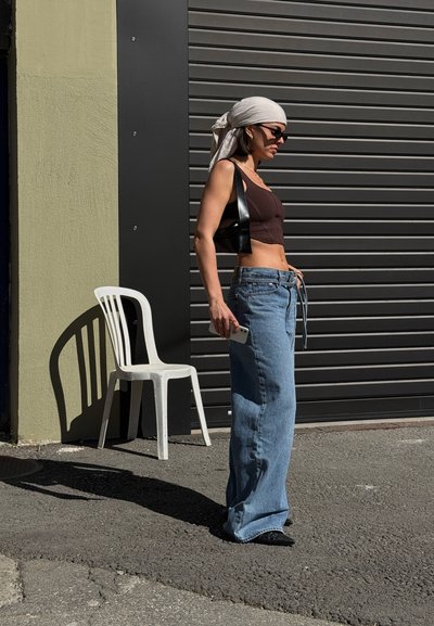 Jeans de denim azul de pierna ancha con cintura alta, combinados con un top corto marrón. Accesorios incluyen un bolso negro y gafas de sol. Silla blanca sencilla al fondo.