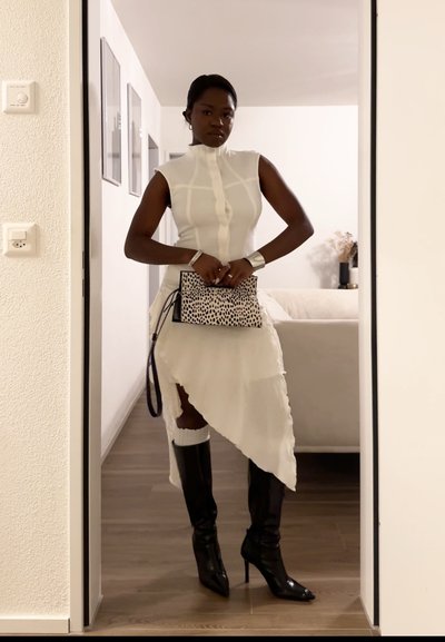 Mujer con vestido asimétrico blanco sin mangas y botas negras hasta la rodilla, sosteniendo un bolso de mano con manchas, de pie en la puerta de una habitación minimalista.