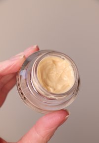 Kreminė beige drėkinamoji priemonė skaidriame stiklo inde. Matoma lygi tekstūra, su plastiku dangteliu. Rankos laiko indą, rodančią pirštų detales.