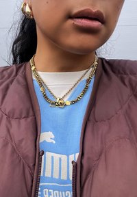 Joalharia em metal dourado com múltiplas correntes, apresentando pendentes circulares. Colete acolchoado castanho escuro por cima de um sweatshirt azul com gráficos brancos.