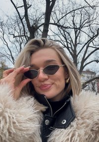 Elegantna sončna očala v obliki srca s senčenimi črnimi lečami in z zlatimi poudarki. Model nosi črno usnjeno jakno, obloženo z belo krzno.