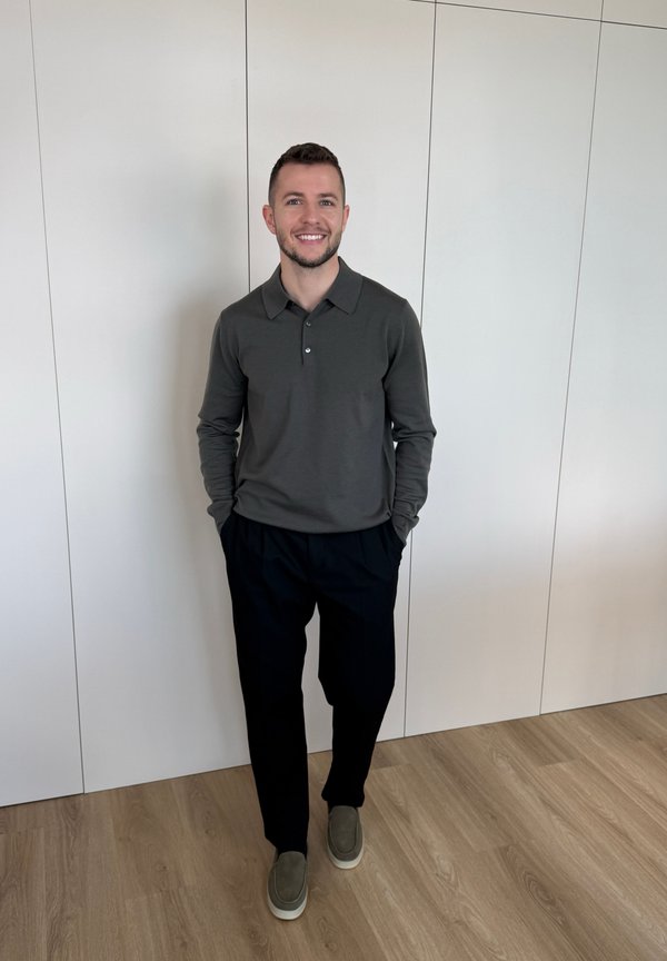 Muž stojí vnútri pri bielej stene, má na sebe tmavé polokošeľu s dlhým rukávom, čierne nohavice a sivé slip-on topánky, usmieva sa s rukami v kapsách.