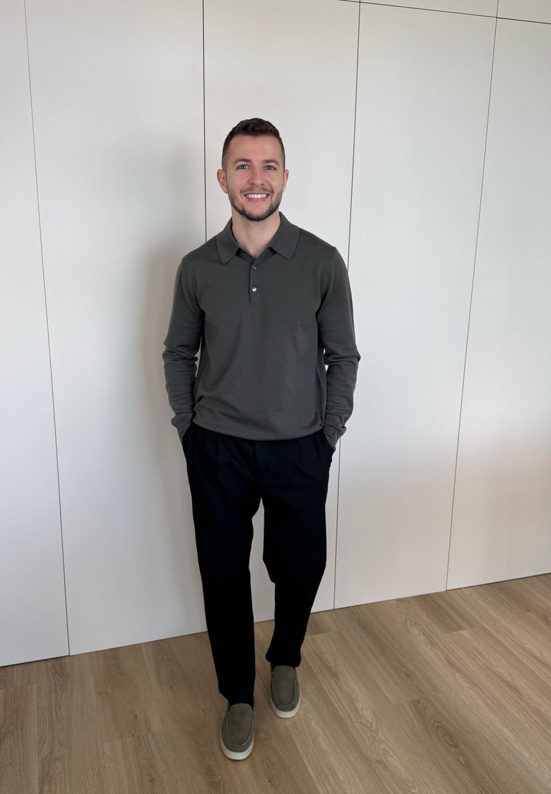 Άνδρας στέκεται μέσα σε ένα δωμάτιο μπροστά σε λευκό τοίχο, φοράει σκούρο πόλο με μακριά μανίκια, μαύρο παντελόνι και γκρι παπούτσια χωρίς κορδόνια, χαμογελά με τα χέρια στις τσέπες.