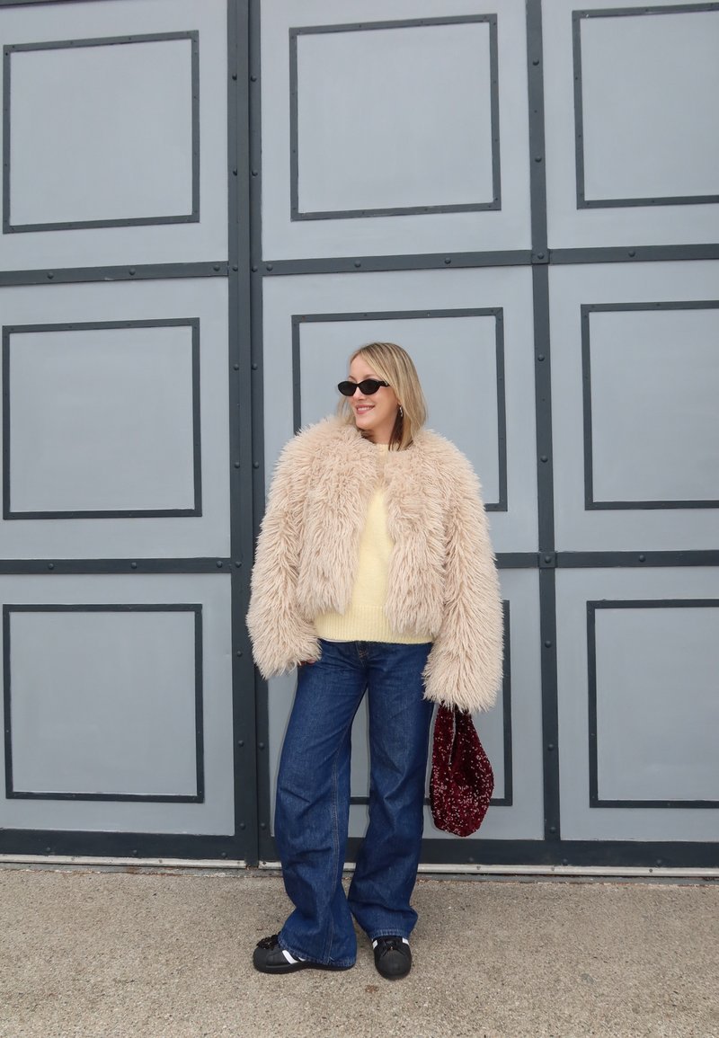 Veste en fausse fourrure beige, pull jaune, jean bleu à jambes larges, chaussures noires et sac à main texturé bordeaux sur un fond de panneaux gris.