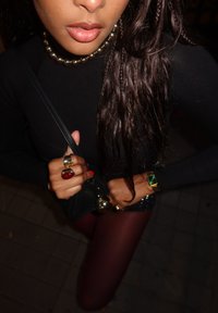 Μαύρη μπλούζα με μακριά μανίκια. Λαμπερό κολιέ από μαργαρίτες, πολλαπλοί δακτύλιοι με κόκκινες και πράσινες λεπτομέρειες, κρατάει μια μικρή μαύρη τσάντα. Σκούρες κολάν με γυαλάδα.