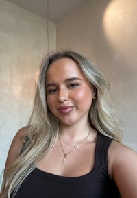 Kvinde med langt, bølget blondt hår iført en brun ærmeløs top og lagdelte gyldne halskæder med dekorative vedhæng mod en lys baggrund.