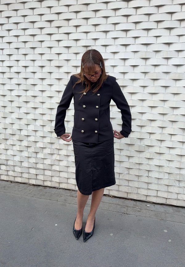 Femme portant une veste noire à double boutonnage, une jupe noire et des talons noirs, debout, regardant vers le bas et ajustant ses manches contre un mur blanc texturé.