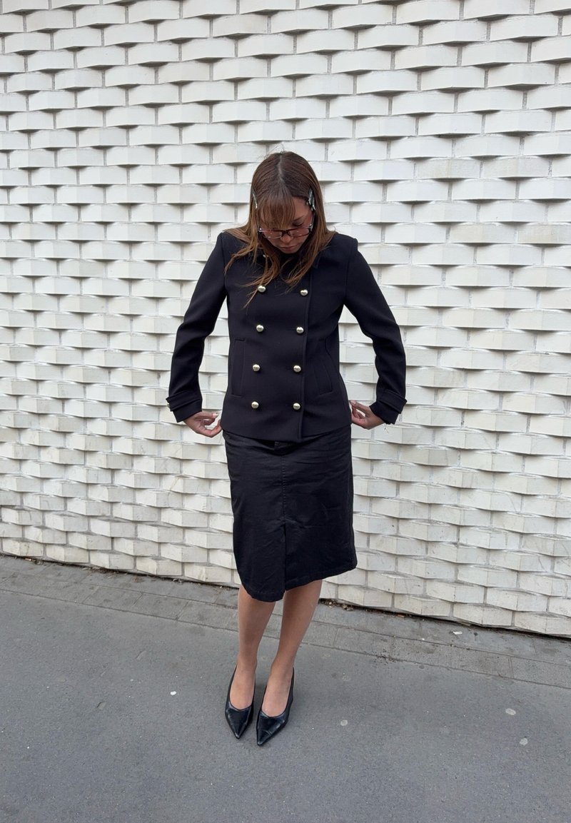 Femme portant une veste noire croisée, une jupe noire et des talons noirs, debout, regardant vers le bas et ajustant les manches contre un mur blanc texturé.