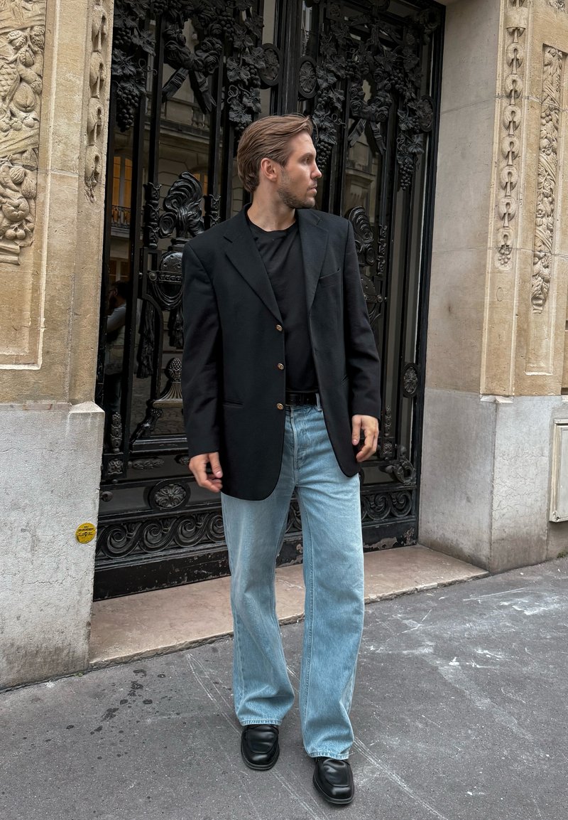 Blazer nero con bottoni dorati, t-shirt nera, jeans a gamba larga azzurri chiari e scarpe nere, su sfondo di un'imponente cancellata in ferro nero decorata.
