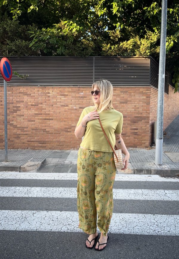 Vrouw met zonnebril en groene outfit staat op een zebrapad, met een gevlochten tas in haar hand, met op de achtergrond een bakstenen muur en bomen.