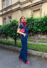 Denim jumpsuit i mørkeblå med brede ben og lomme detaljer, parret med en rød mønstret håndtaske og matchende røde hæle. Grønne planter i baggrunden.