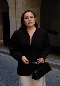 Czarny strukturalny żakiet na białej sukience, trzymając okulary przeciwsłoneczne i czarną torebkę. Gładka tkanina, fason dopasowany do sylwetki i minimalistyczne akcenty.